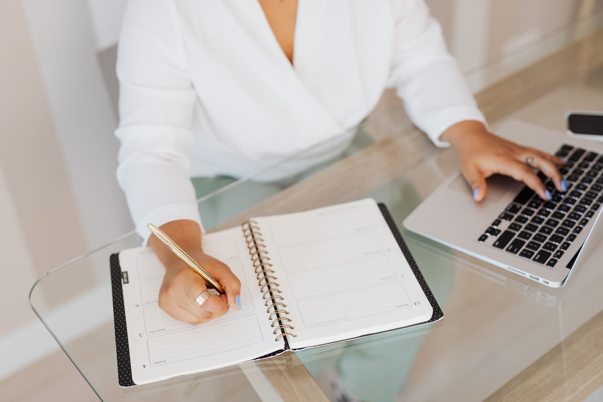 A person writing in a planner notebook beside an open laptop on a glass desk